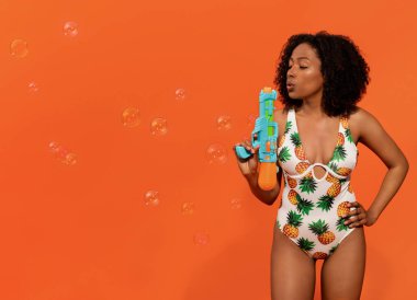 Şirin, neşeli, şık mayo giyen, sabun köpüklerinin arasında su tabancası tutan, turuncu stüdyo arka planında eğlenen, fotokopi çeken güzel siyah bir kadın. Yaz tatilinde su aktivitesi