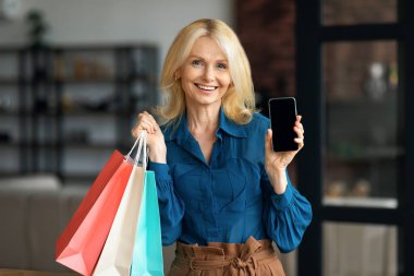 Gülümseyen olgun bir kadın boş akıllı telefon gösteriyor ve elinde parlak alışveriş torbaları, ev içi modeller tutuyor. İnternetten satın alma uygulaması öneren mutlu kadın.