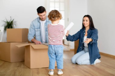 Yeni eve taşındıktan sonra aile, karton kutuları boşaltıyor, mutlu genç ebeveynler ve küçük oğulları, apartman dairesine taşındıktan sonra eşyalarını oturma odasına yerleştiriyorlar.