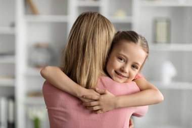 Anneler Günü Konsepti. Neşeli küçük kız, evde annesine sımsıkı sarılıyor, gülümseyen kadın çocuğu kucaklıyor, mutlu aile, oturma odasında birbirine bağlanıyor,