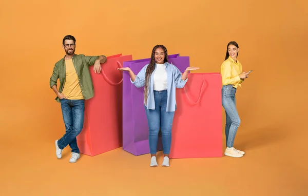 消费主义 季节性销售 黑色星期五的概念 快乐的多种族千禧一代在橙色背景的巨大的彩色购物袋旁 拼凑在一起 — 图库照片