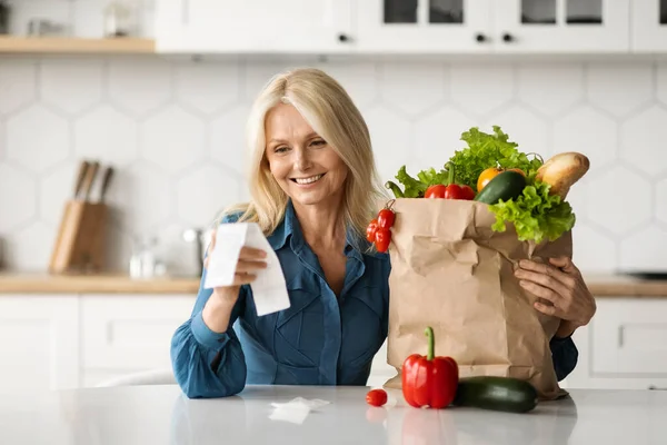 Shopping Economy. Happy Mature Woman In Kitchen Holding Bag Of Groceries And Checking Bill, Beautiful Elderly Female Sitting At Table And Counting Spends, Enjoying Affordable Prices For Food