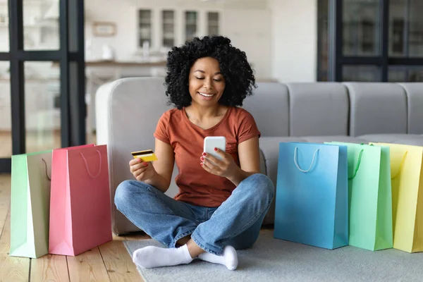 オンラインショッピングコンセプト 明るい買い物袋の近くの床に座っている間 自宅でスマートフォンやクレジットカードを使用して幸せな黒人女性 笑顔アフリカ系アメリカ人女性インターネット購入を行う — ストック写真