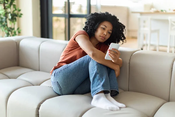 沮丧的非洲裔美国女士坐在沙发上看着手机屏幕 心情烦躁不安 她收到了坏消息 看到了令人不快的信息 或在等待重要的电话 抄袭了一个空格 — 图库照片
