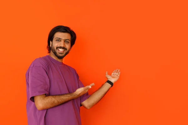 紫色のTシャツを着た幸せなハンサムな若いインド人男性は オレンジのスタジオの背景に隔離された広告や笑顔のためのコピースペースを指しています 素晴らしいオファー 偉大な契約の概念 — ストック写真