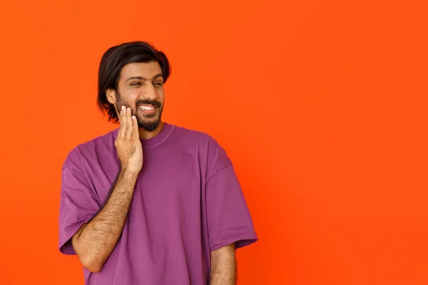 紫のTシャツを着た懐疑的な笑顔の若いハンサムな東の男は オレンジのスタジオの背景にコピースペースを見ています 広告を見ながら顔に触れるインド人男性 — ストック写真