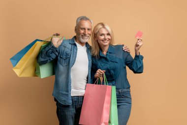 Renkli alışveriş çantalarıyla poz veren kot pantolon giymiş, bej renkli stüdyo arka planda banka kartı gösteren mutlu ve güzel yaşlı bir çift. Sınırsız ödeme, sınırsız alışveriş.