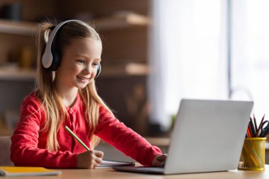 E-Öğrenme Konsepti. Gülümseyen, kulaklıklı, evde dizüstü bilgisayarla çalışan, bilgisayar ekranına bakan, notlar alan, internetten ders alan, interneti kopyalayan genç bir kız.