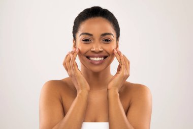 Yüzüne masaj yapan, gülümseyen, pastel renkli arka planda soyutlanmış binlerce yıllık neşeli siyah kadın. Yaşlanma önleyici cilt bakımı, yüz gerdirme masajı