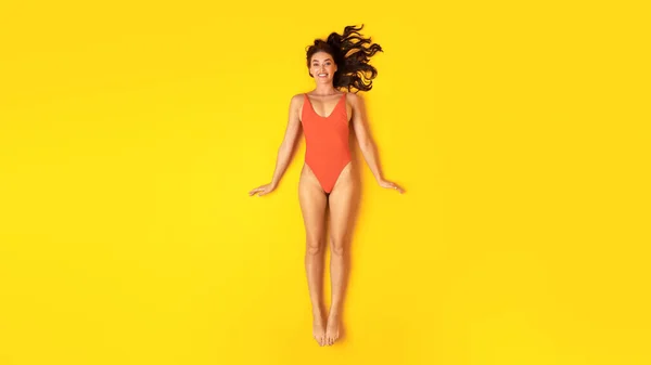 夏休みの準備 黄色のスタジオで寝そべっているカメラに笑顔を投げかけるオレンジ水着のハッピーレディ背景 パノラマ 上からの眺め スリム女性の日光浴と水着を着てリラックス — ストック写真