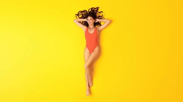 夏休み オレンジ色のワンピース水着を着た若い女が日光浴をして黄色いスタジオの背景でリラックスし カメラに微笑む 水着姿の女性のトップビュー撮影 パノラマ — ストック写真