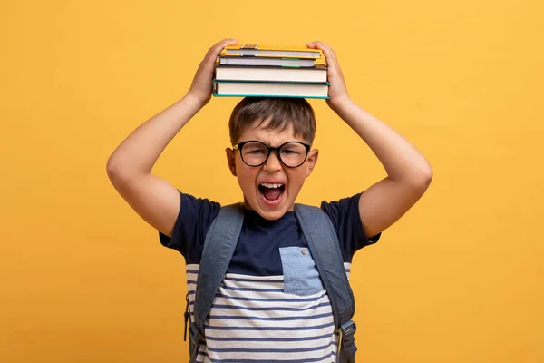 バックパックを持って眼鏡をかけ 彼の頭の上に本を身に着けているかわいい男の子の学校を強調悲鳴 学校での困難の苦しみ 黄色のスタジオの背景に隔離 — ストック写真