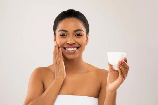 Uśmiechnięta Młoda Pogodna Czarna Kobieta Owinięta Ręcznik Białym Słoikiem Kosmetykami — Zdjęcie stockowe