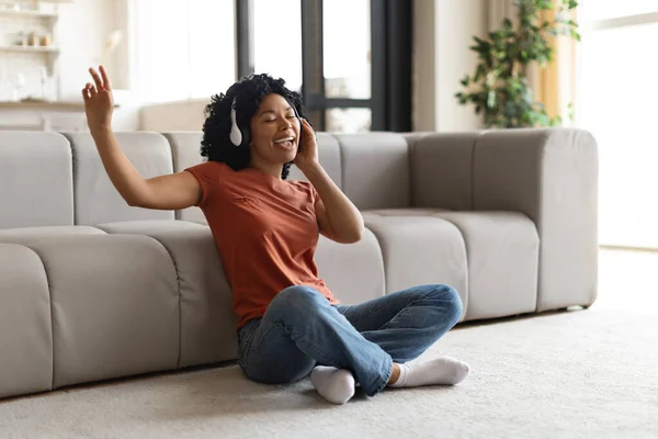 国内の楽しみ 自宅でワイヤレスヘッドフォンで音楽を聴いている陽気なアフリカ系アメリカ人女性 リビングルームで床に座っている幸せな黒人女性 彼女のお気に入りのプレイリストを楽しみます スペースをコピー — ストック写真