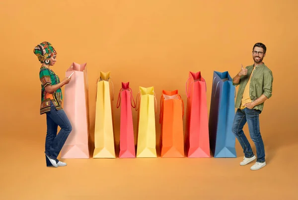 快乐美丽的混血夫妇在网上购物 黑人女士和留着胡子的高加索男人站在巨大的彩色购物袋旁 用手机 用橙色背景展示大拇指 拼凑在一起 — 图库照片