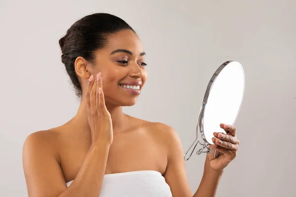 スキンケアルーチン 小さな鏡を見て幸せな魅力的なアフリカ系アメリカ人の千年紀の女性 指と笑顔で顔をマッサージ クリームを適用 血清または油 灰色で隔離された コピースペース — ストック写真