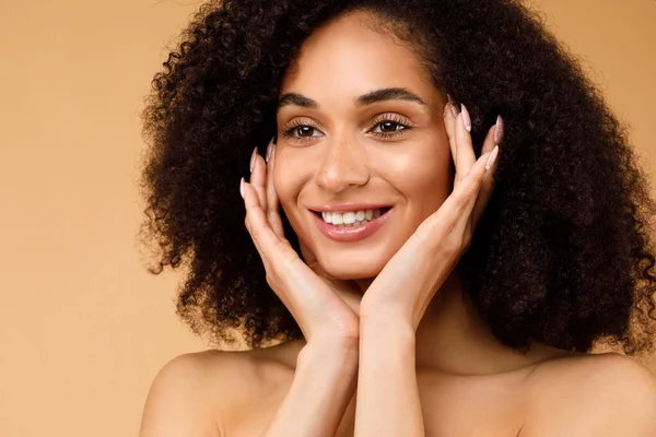 Headshot of happy young black lady touching her cheeks, enjoying silky skin after using luxury organic premium beauty face care products, beige background, closeup