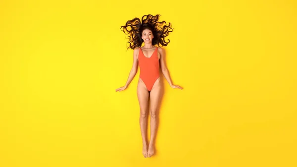 夏の美女 黄色のスタジオの背景に横たわるカメラに笑顔オレンジ水着でかなりスリム女性 日光浴や休暇中のリラックス パノラマ トップビューショット — ストック写真
