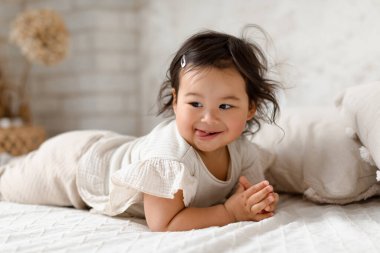 Babyhood. Cute Korean Baby Girl Posing Looking Aside Lying On Stomach On Bed At Cozy Home Interior. Shot Of Funny Little Toddler Wearing Comfortable Clothing Having Fun In Bedroom clipart