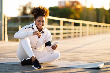 Akıllı telefonlu ve Protein barlı mutlu siyah bayan dışarıda spor yaptıktan sonra dinleniyor spor giyim kuşağında gülümseyen Afro-Amerikan kadın dışarıda spor minderinde oturuyor ve cep telefonu kullanıyor.