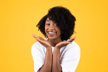 Bin yıllık Afro-Amerikan kıvırcık saçlı beyaz tişörtlü kadının eğlenmesine, boş zamanlarının tadını çıkarmasına sarı stüdyo arka planında izole edilmesine sevindim. Yaşam tarzı, güzellik, sürpriz, reklam ve teklif, öğrenci duyguları.