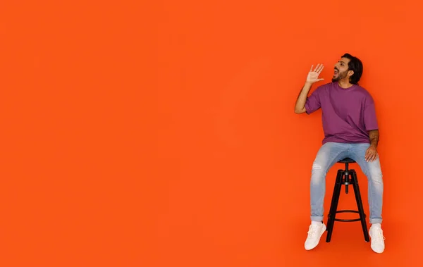 年轻帅气的年轻的东方人穿着时髦的休闲装 坐在橙色工作室的椅子上 对着广告的复制空间大喊 做通告 全长拍摄 — 图库照片