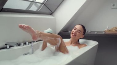 Kendine bakmanın neşesi. Kaygısız beyaz kadın mutlu köpük banyosu ritüelinin tadını çıkarıyor, bacaklarını sünger ve duş jeliyle yıkıyor, çekim yapıyor, boş bir alanda.