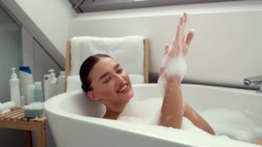 Mutluluk sükuneti. Genç ve mutlu bir bayan evde köpük banyosu yapıyor, ellerini sabun köpüğüyle yıkıyor ve gülümsüyor, yavaş çekim, boş alan.
