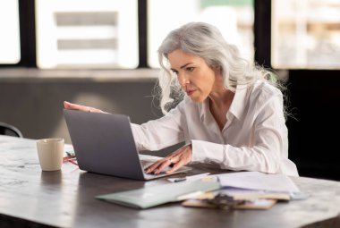 Ofis işleri rutini. Orta yaşlı iş kadını dizüstü bilgisayarda çalışıyor, iş yerinde zor görevleri yazıyor ve çözüyor, kapalı mekanda masa başında oturuyor.