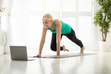 Çevrimiçi Fitness. Evde dizüstü bilgisayarın önünde egzersiz yapan sportif olgun bir bayan. Aktif son sınıf öğrencisi. Video dersleri izleyip spor yapıyor.