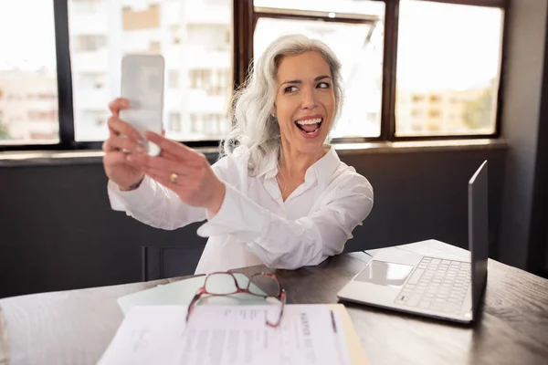 Workplace Fun. Joyful Middle Aged Businesswoman Making Selfie On Phone In Modern Office, Sitting At Table With Laptop And Celebrating Career Growth And Business Success. Selective Focus