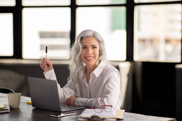 Business Leadership. Successful Manager Woman Sitting Near Laptop, Posing At Workplace Holding Pen And Smiling To Camera Indoor. Happy Businesswoman Working Online In Modern Office