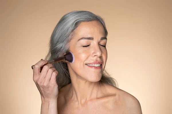 粉を適用する灰色の髪を持つ積極的なシニア女性 彼女の顔にブラシで赤面し ベージュの背景に孤立ポーズ アンチエイジング化粧品やヌードメイク 美容ケア 広告やオファー — ストック写真