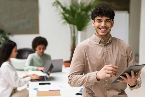 Digital Business Offer. Joyful Middle Eastern Businessman With Tablet Standing At Corporate Meeting In Modern Office, Posing In Front Of Colleagues And Smiling To Camera. Career And Leadership