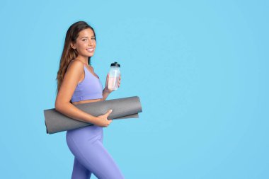 Bin yıllık beyaz kadın spor giyimli, minder ve su şişesi hazır, mavi stüdyo arka planında soyutlanmış. Spor, kilo kaybı ve vücut bakımı, yoga, su dengesi