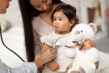 Bebek Sağlığı Kontrolü. Pediatrist Bayan Stetoskop kullanıyor. Klinikteki randevusu sırasında Asyalı Çocuk Oyuncak Kalp atışlarını dinliyor. Anne ve Kızı Aile Doktorunu Ziyaret Ediyor