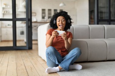 Evde akıllı telefonda video oyunu oynayan heyecanlı siyahi kadın, mutlu Afrika kökenli Amerikalı kadın cep telefonu saklama ve oturma odasında otururken online zaferi kutlayan,