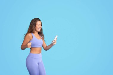 Bin yıllık beyaz kadın spor giyimli, akıllı telefonlu, mavi stüdyo arka planında izole bir şekilde eğleniyor. Spor uygulaması, kilo verme ve vücut bakımı için blog, iyi haber ve sıçrama