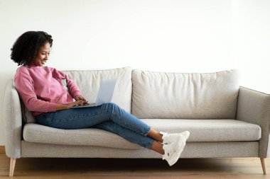 Bin yıllık Afro-Amerikan kıvırcık kadın dizüstü bilgisayarda yazı yazıyor, oturma odasındaki kanepede sohbet ediyor. Ders, video iletişimi, evde çalışmak ve çalışmak.