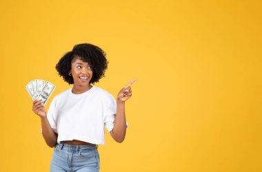Beyaz tişörtlü Afro-Amerikan kıvırcık saçlı mutlu bir milenyum kadını yüklü miktarda para tutar, boş alanı işaret eder, sarı stüdyo arka planında izole edilir. Reklam ve teklif, kâr, mali tasarruflar