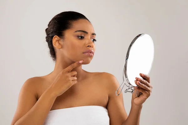 Skin disorders. Unhappy sad young beautiful half-naked black woman looking at mirror, african american woman touching skin and touching pimples on her chin skin, studio background