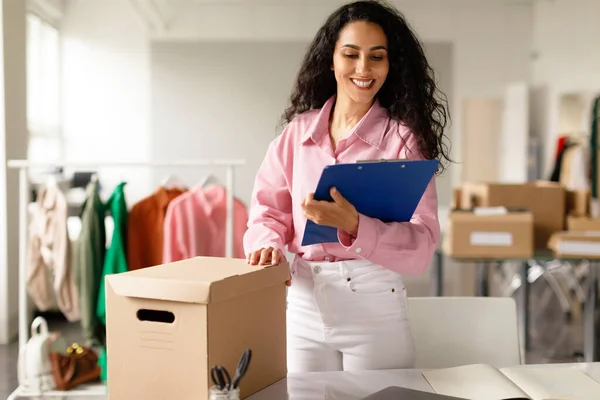 Retail Business. Successful Businesswoman In Fashion Clothing Store Holding Clipboard, Taking Notes Standing Near Table With Cardboard Boxes With Clothes In Showroom, Ready For Delivery To Buyers