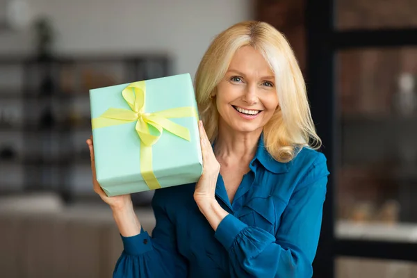 stock image Portrait of happy mature woman sitting on couch at home, holding wrapped gift box, looking and smiling at camera, female receiving birtday or anniversary present, copy space