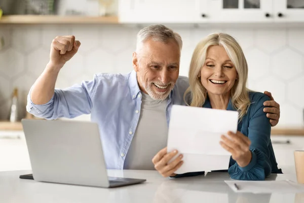 Portrait of excited senior spouses with papers celebrating good news in kitchen, happy mature couple reading mail and enjoying success, emotionally reacting to credit approvance or financial profit