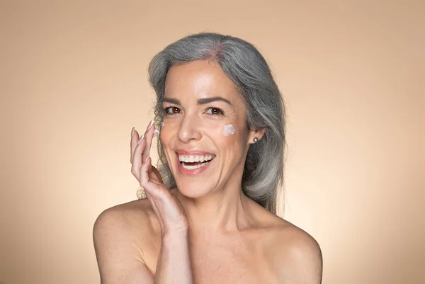 高齢女性の顔のスキンケア 頬や笑顔に保湿クリームを適用ハッピーシニア女性 保湿肌 ベージュスタジオの背景にポーズ フリースペース — ストック写真