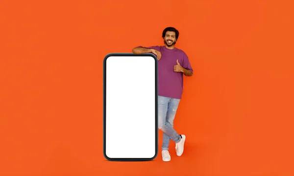 明るくハンサムなひげを生やした若いインド人男性が白い空白の画面で巨大なスマートフォンで立って 親指を示す オンラインオファーやモバイルアプリの広告のためのモックアップ オレンジの背景に隔離された コピースペース — ストック写真