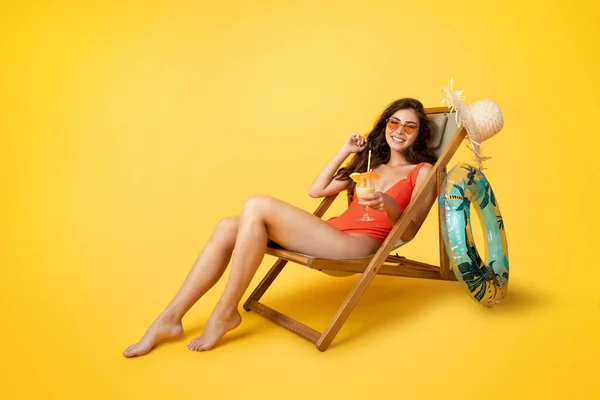 Glad millennial mixed race lady in swimsuit sits in deck chair with glass of tropical cocktail, enjoy vacation, isolated on yellow studio background. Rest, relax, holiday trip in summer