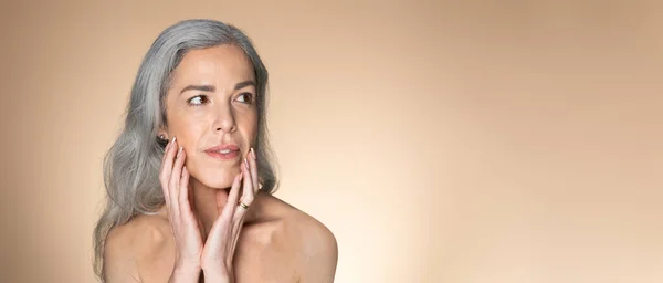 Reflections of time. Grey-haired senior woman touching her glowing face skin and looking aside at copy space, beige studio background, panorama with free space, web-banner