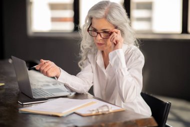 Çevrimiçi Çalışma Programı. Orta yaşlı iş kadını, diz üstü bilgisayara gözlüklerle bakıyor, internete bakıyor ve modern ofiste bilgisayarla çalışıyor. Girişimcilik Kariyeri