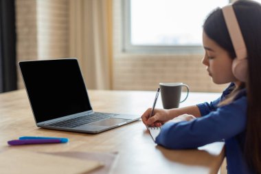 Online Öğrenme Teklifi. Kulaklıklı liseli kız laptopta boş ekranlı notlar alıyor ve ödevlerini evde yapıyor. E-Öğrenme, Modern Okul Eğitimi. Yan Görünüm Görüntüsü, Mockup